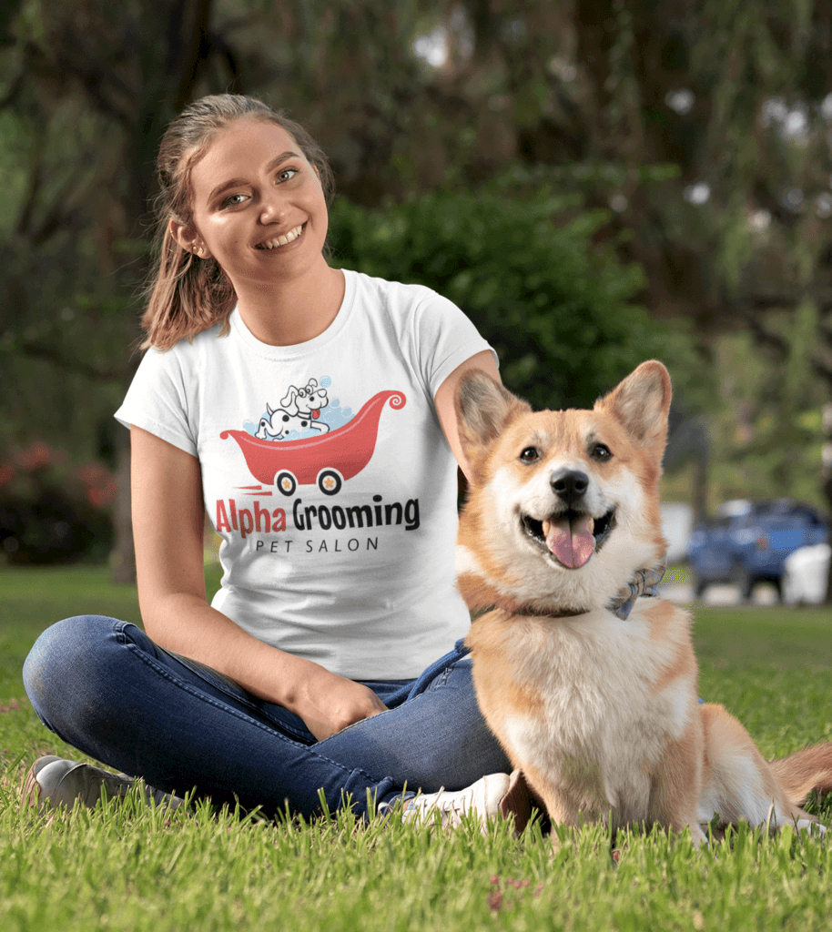 Woman sitting on grass with a smiling dog, wearing a white "Alpha Grooming Pet Salon" t-shirt; park setting with trees in the background—perfectly capturing the joy of mobile pet grooming.